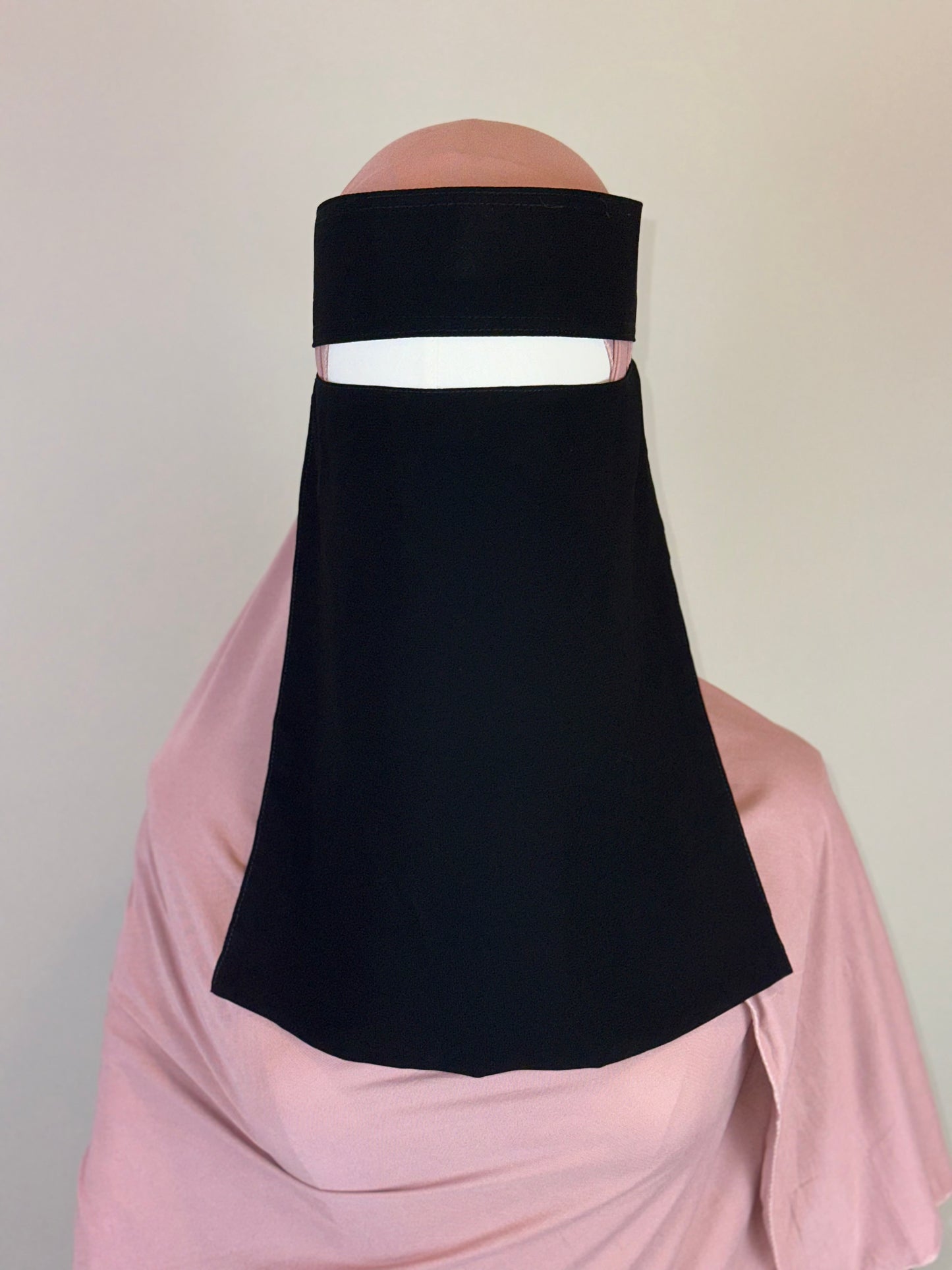 Black niqab veil styled over a light pink hijab on a mannequin against plain background
