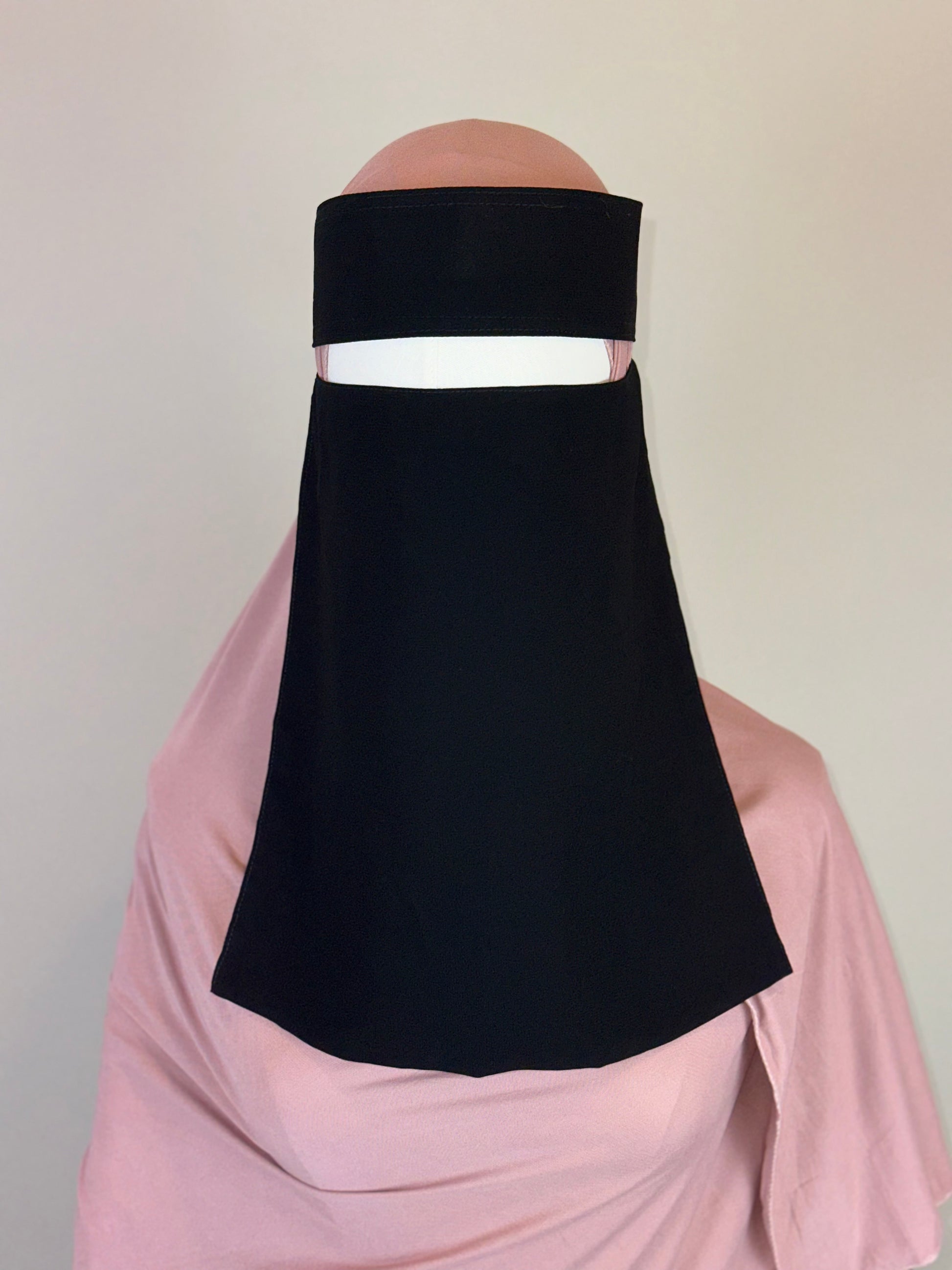 Black niqab veil styled over a light pink hijab on a mannequin against plain background