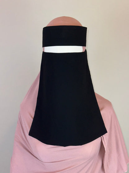 Black niqab veil styled over a light pink hijab on a mannequin against plain background