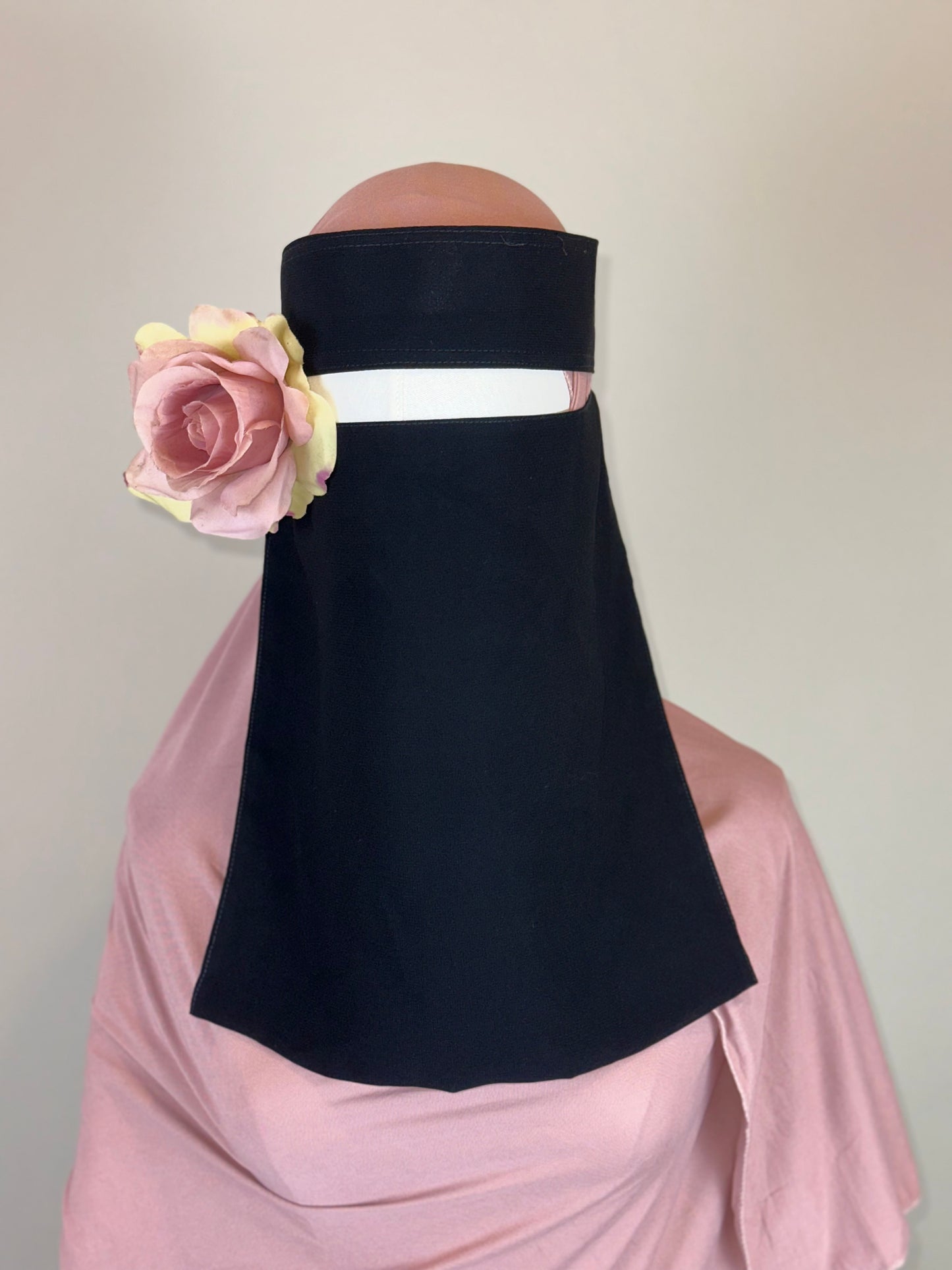 Woman wearing black niqab with pink rose accessory and pink hijab, neutral background