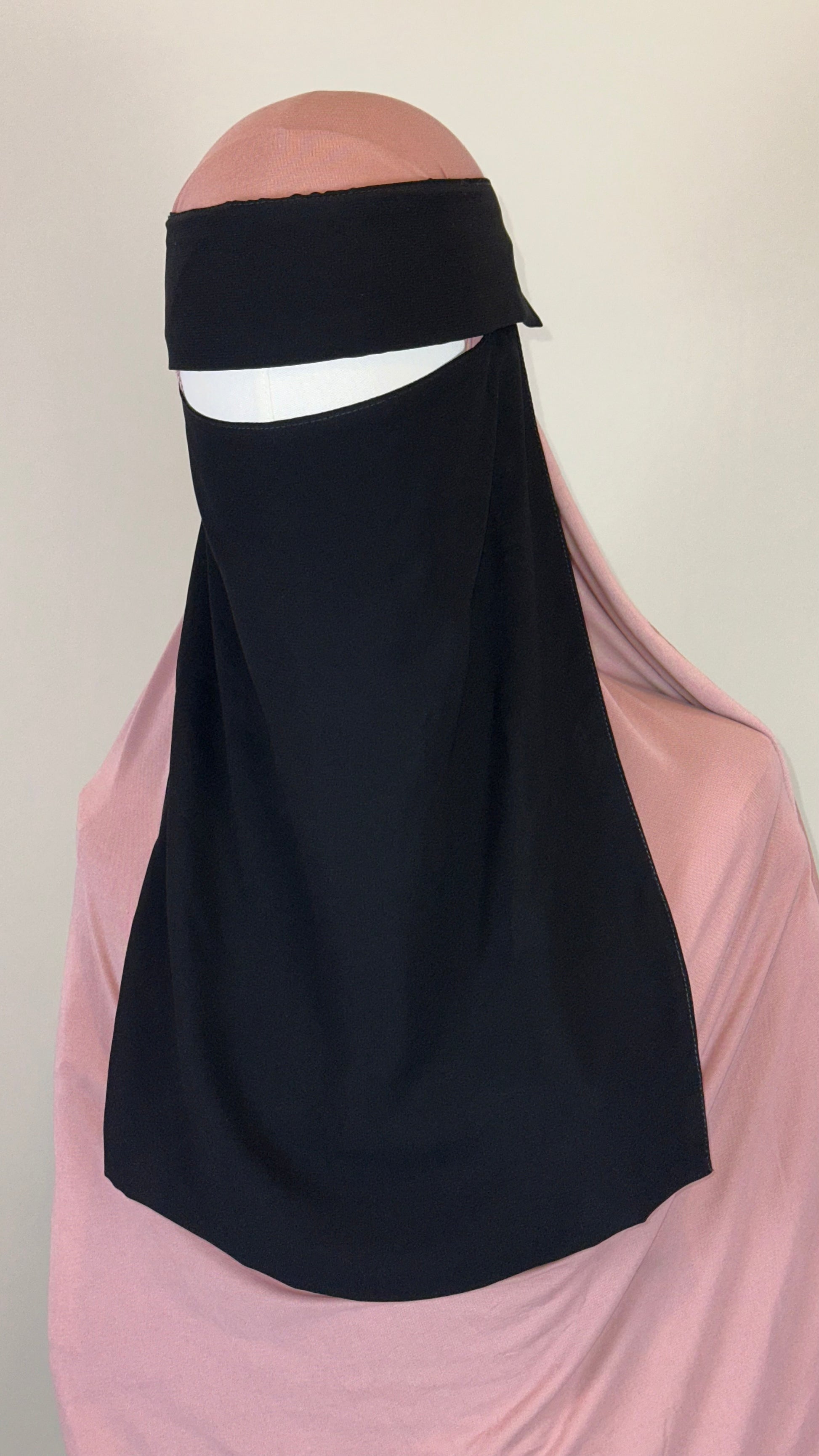 Black niqab over light pink hijab on mannequin against plain background