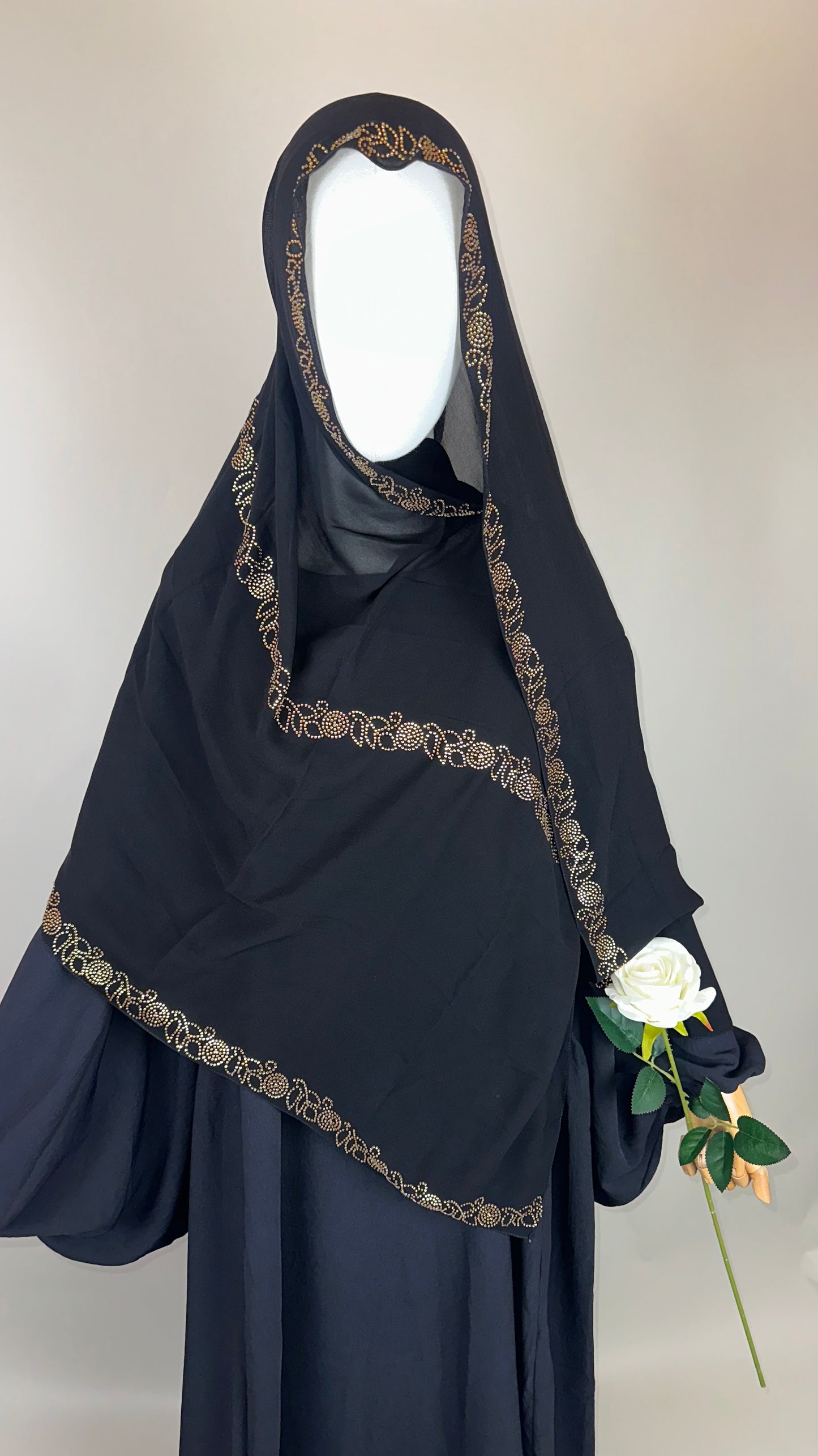 Black embroidered hijab with gold trim, mannequin holding white rose, neutral background